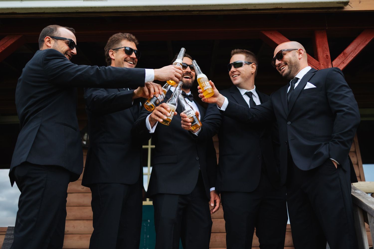Groomsmen in black suits toast with drinks at an outdoor wedding.
