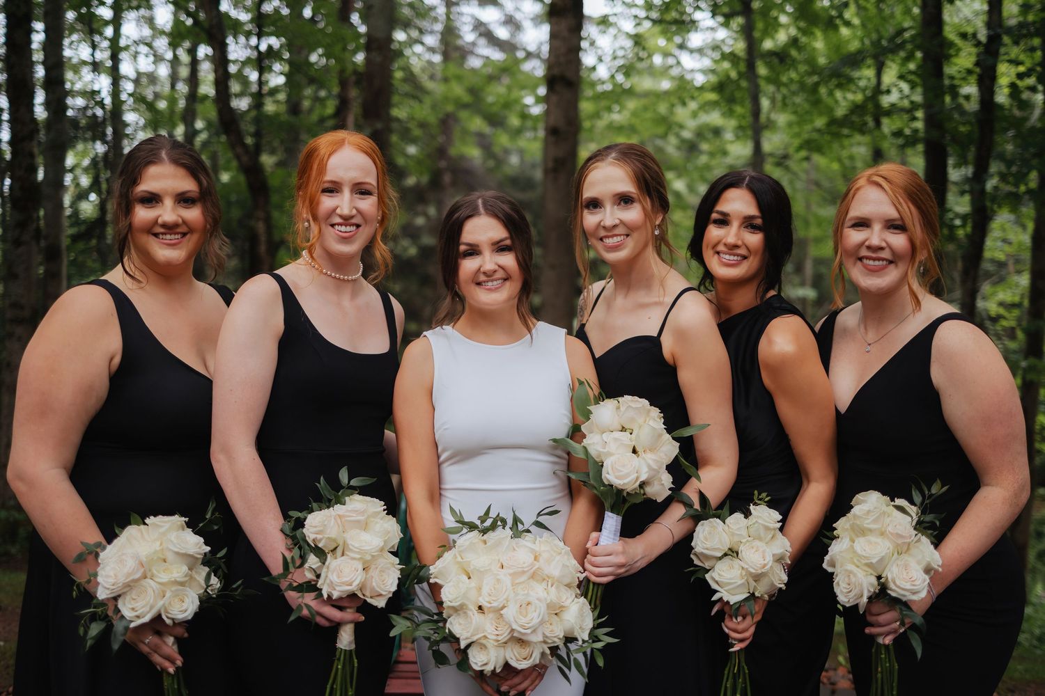 Bridesmaids in black dresses pose with white bouquets against a forest backdrop.