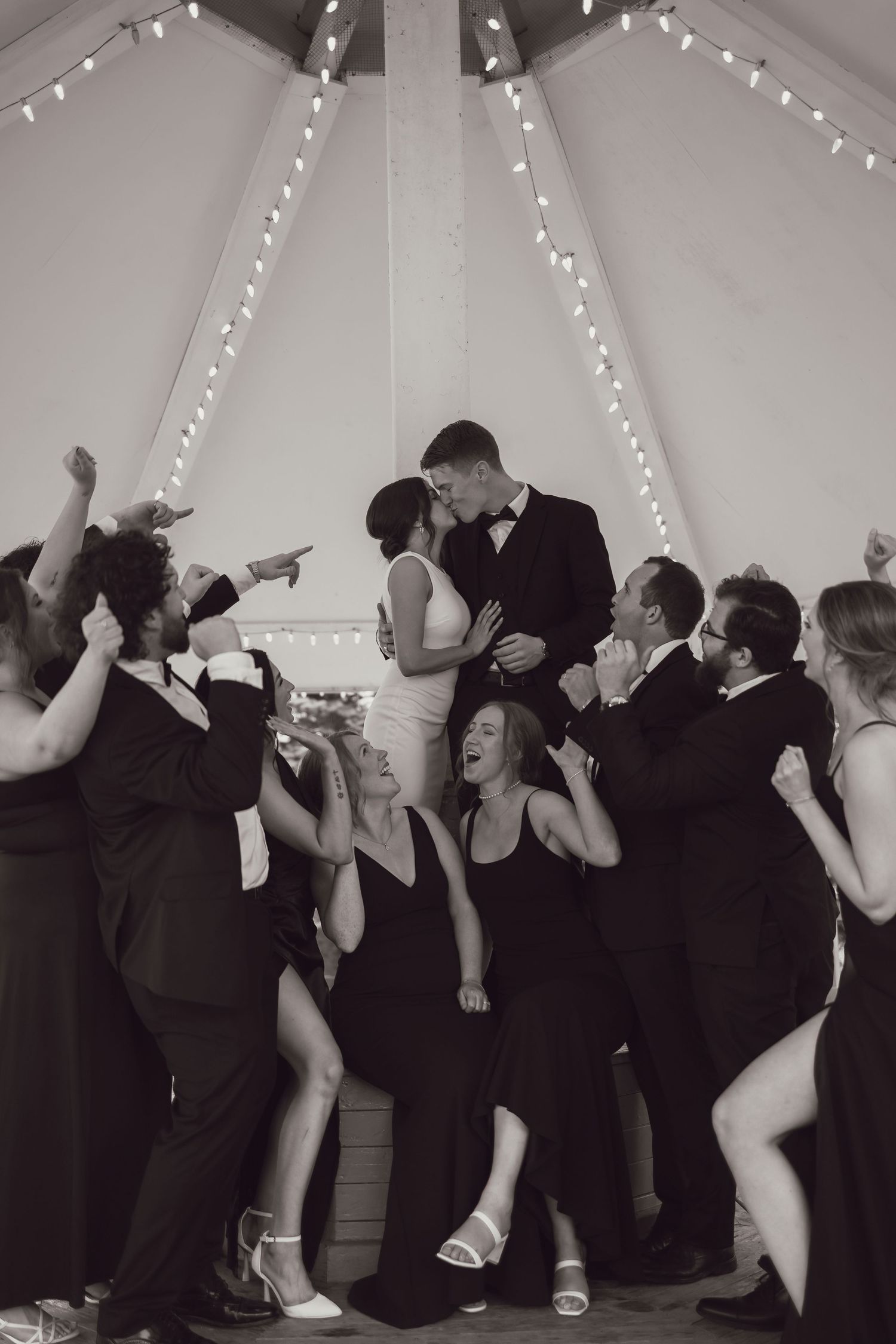 Black and white photo of newlyweds kissing surrounded by celebrating wedding guests under string lights.