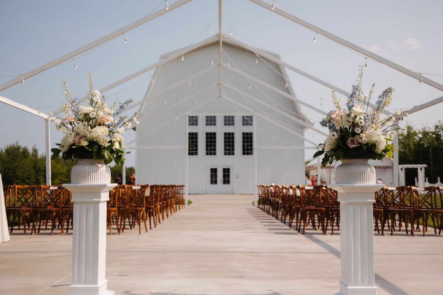 White wedding ceremony tent setup with wooden chairs and floral arrangements.