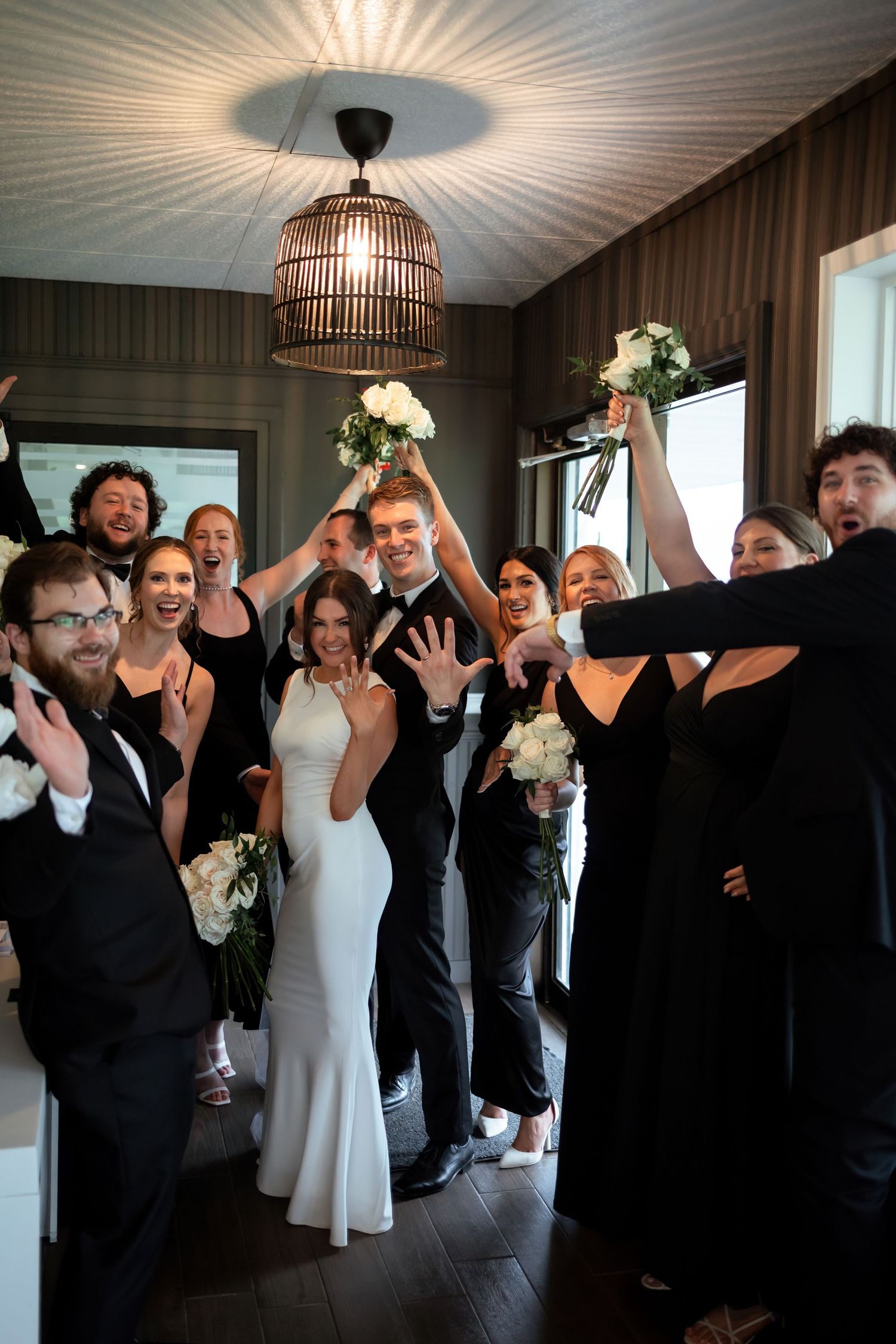 Wedding party celebrates with raised bouquets and hands under a dramatic ceiling light fixture.