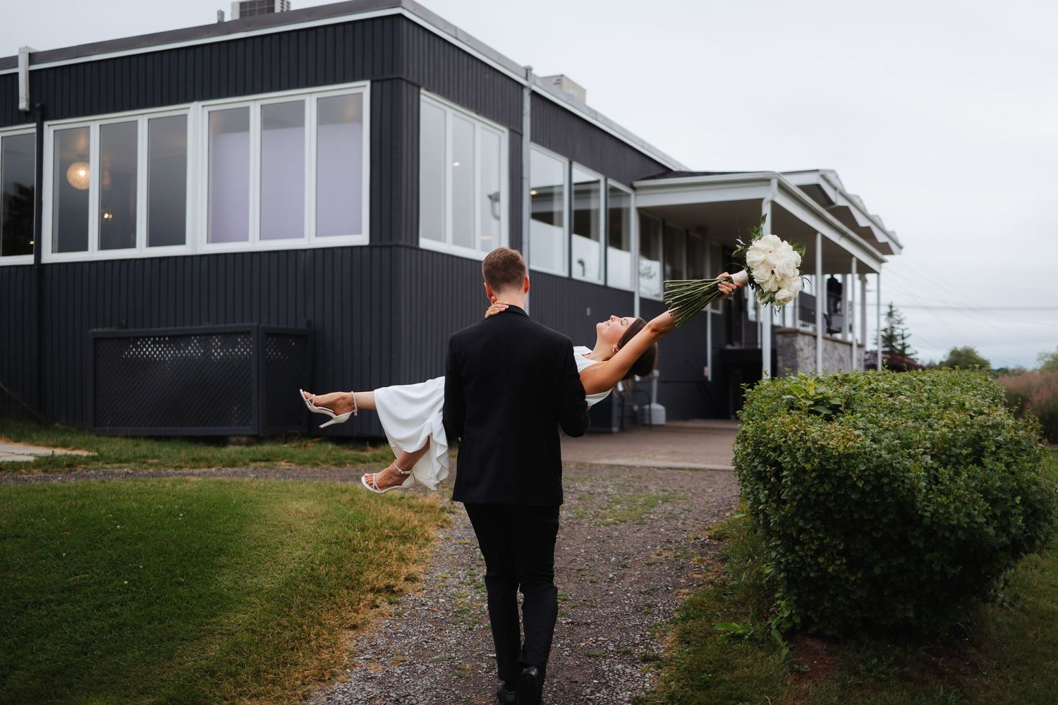 Groom carries bride while walking towards a modern black house exterior.