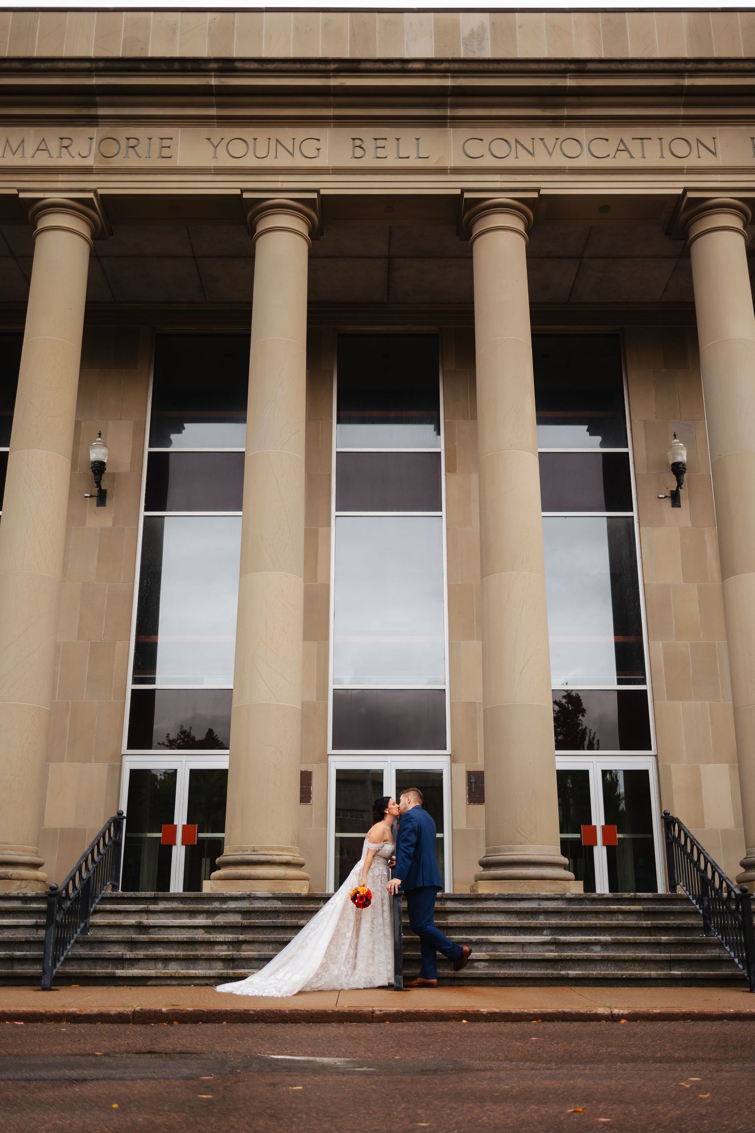 A wedding couple stands between elegant white columns of a historic building's entrance.