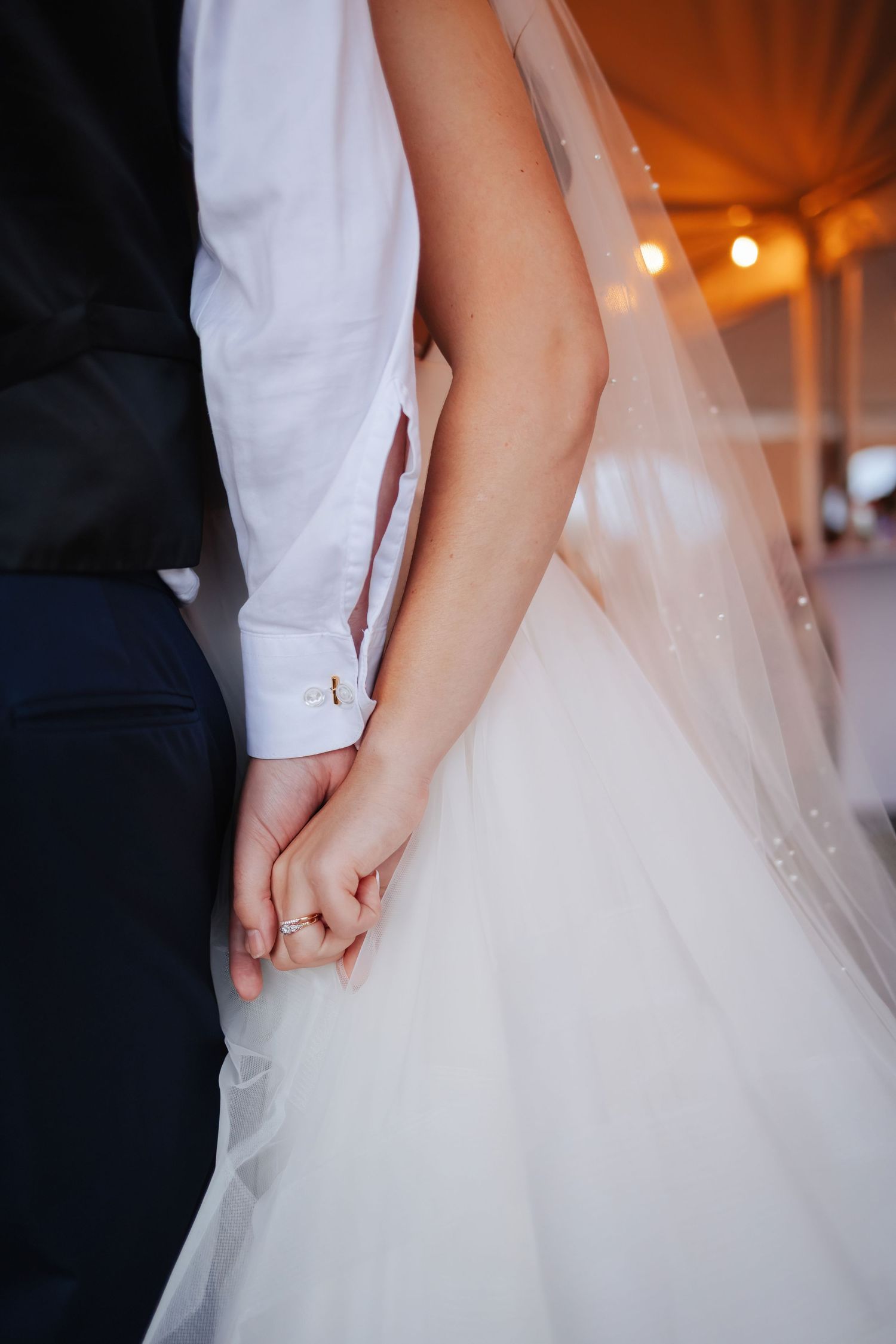 Close-up detail shot of bride and groom holding hands, showing white dress and formal attire.