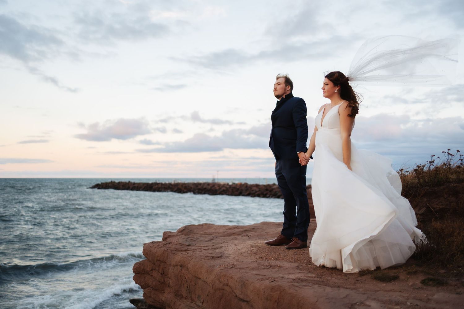 A couple in wedding attire stands on a rocky oceanside cliff during sunset.