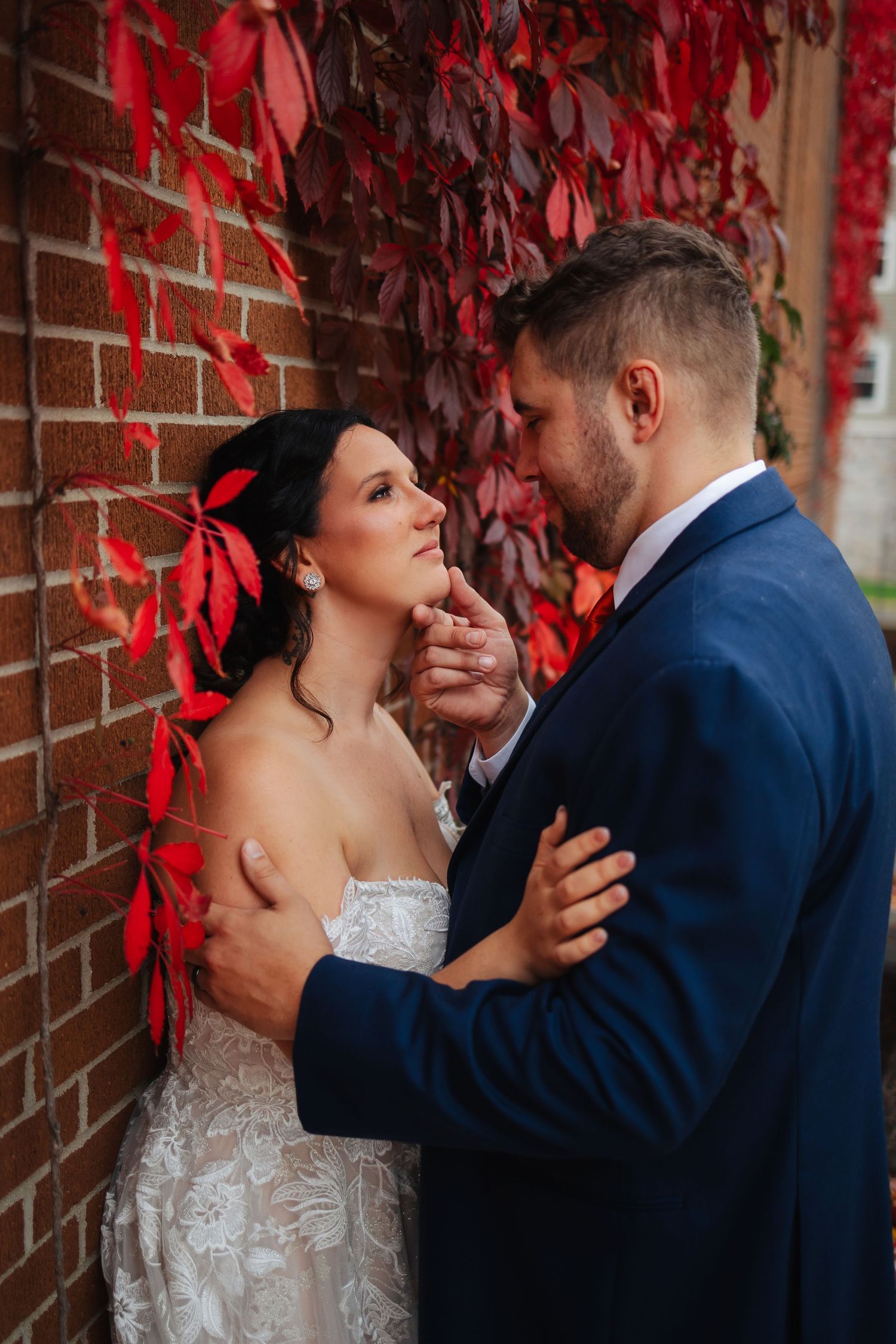 Intimate wedding moment captured under red autumn leaves against a brick wall.