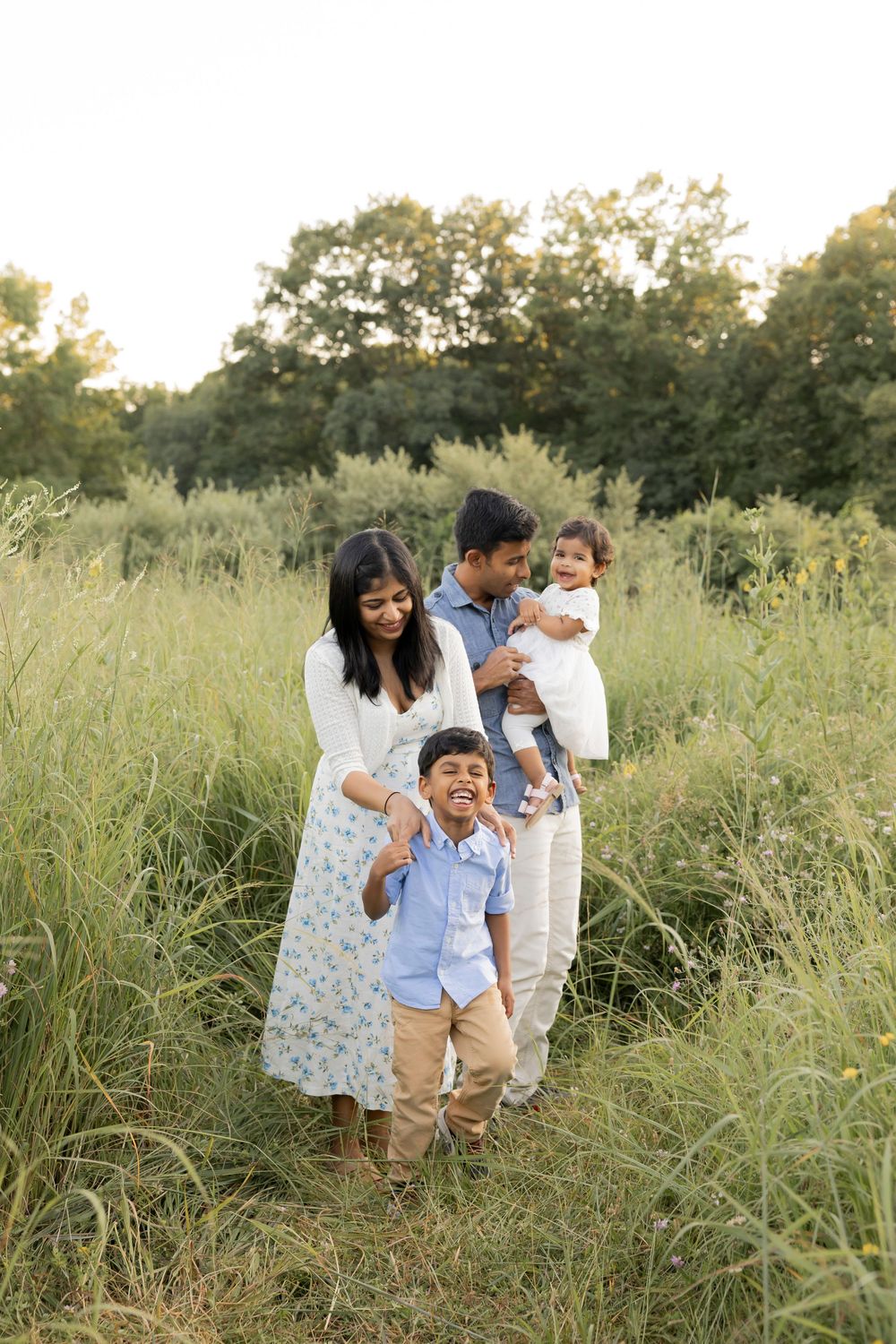 A family playing together through a sunlit meadow with tall grass while wearing blue outfits during golden hour at stoney creek Metropark.