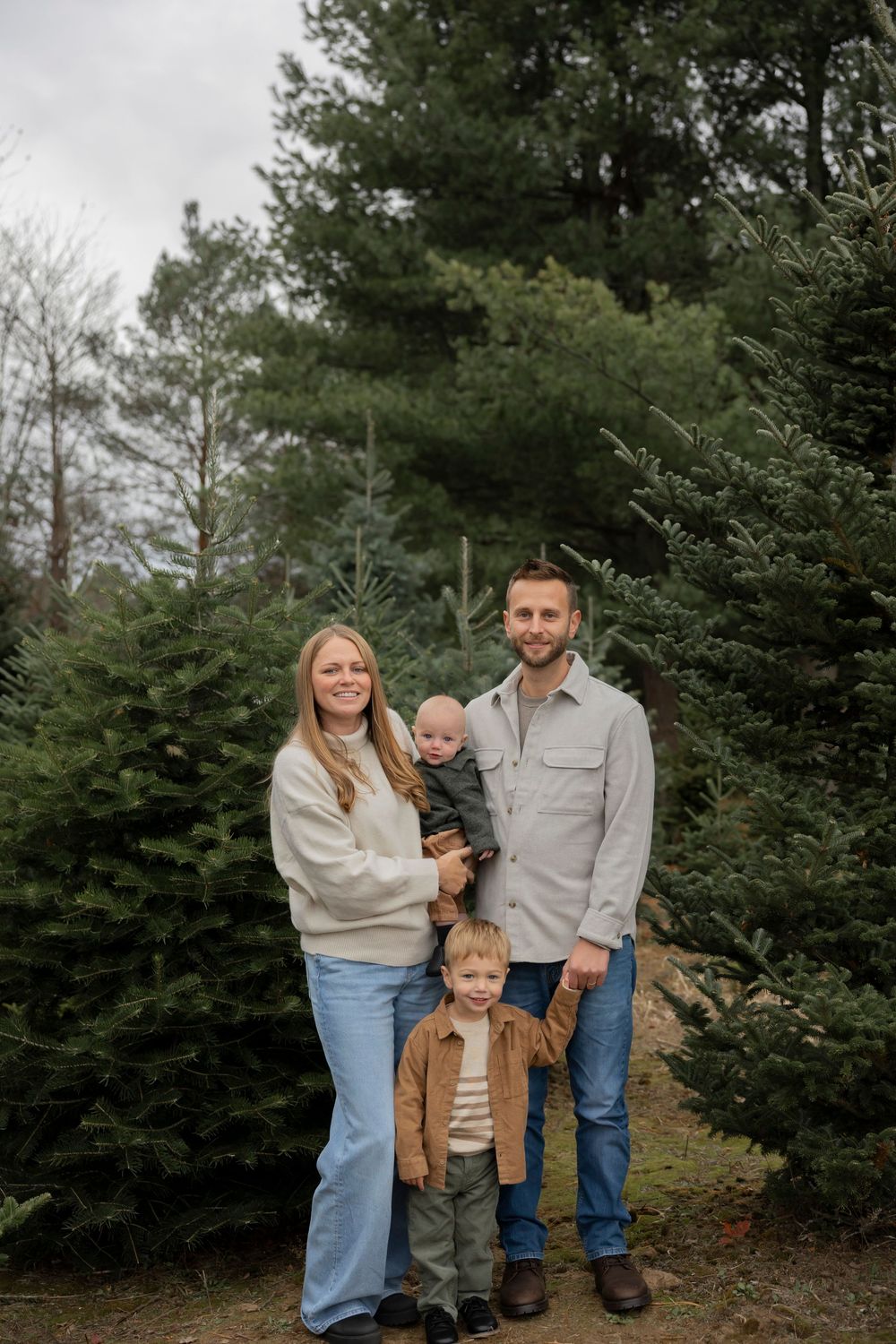 A family stands together among evergreen trees at what appears to be a Christmas tree farm in Lake Orion Michigan on a winter day.