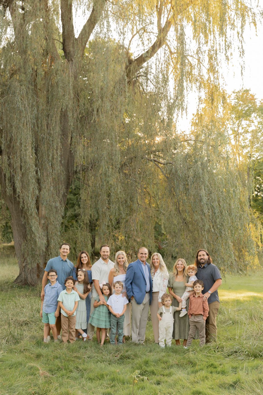 large family dressing in neutrals and pastels all smiling for a photo in front of a large willow tree