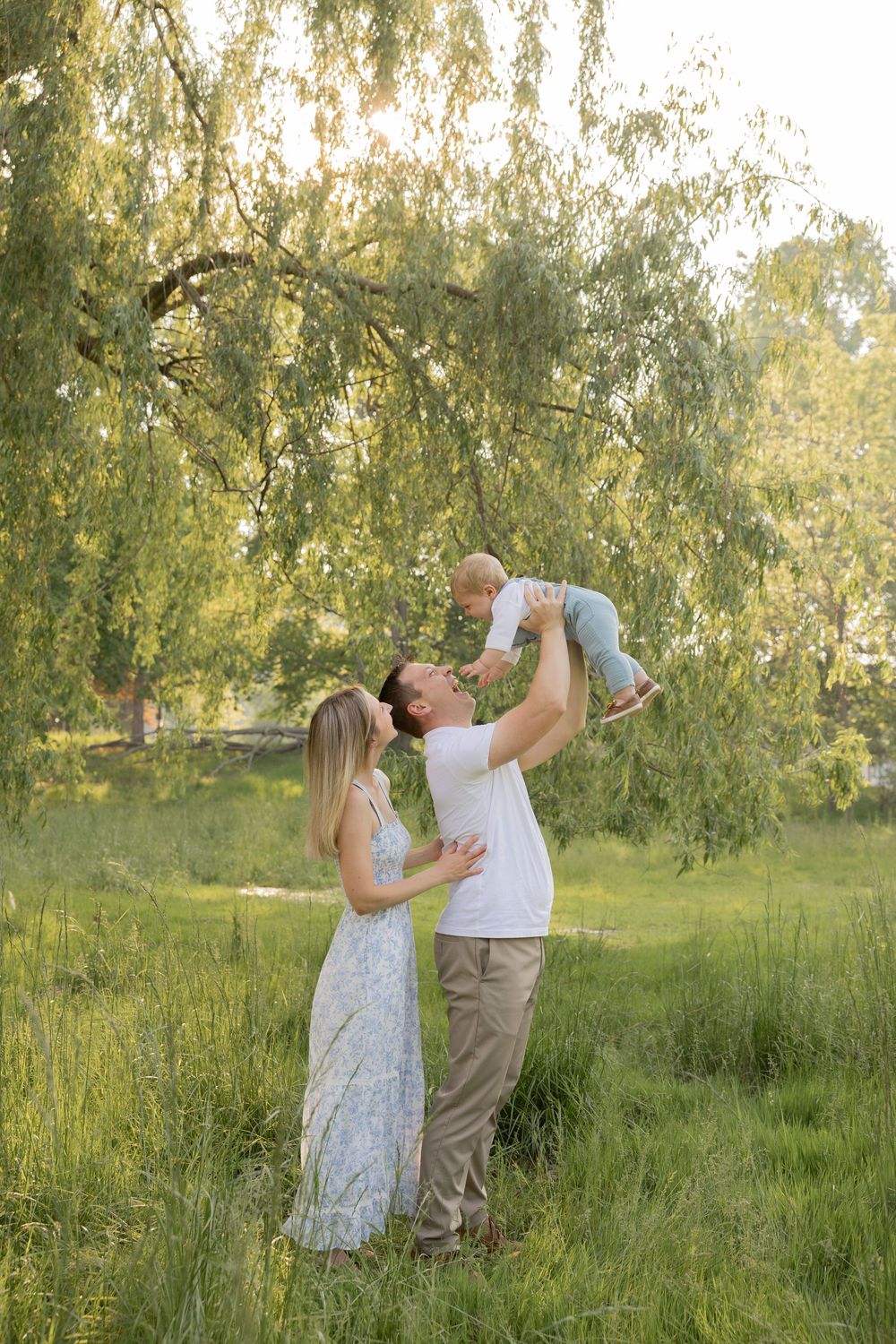 A couple plays airplane with their son in a sunlit meadow with willow trees swaying in the background while they wear blue in Michigan
