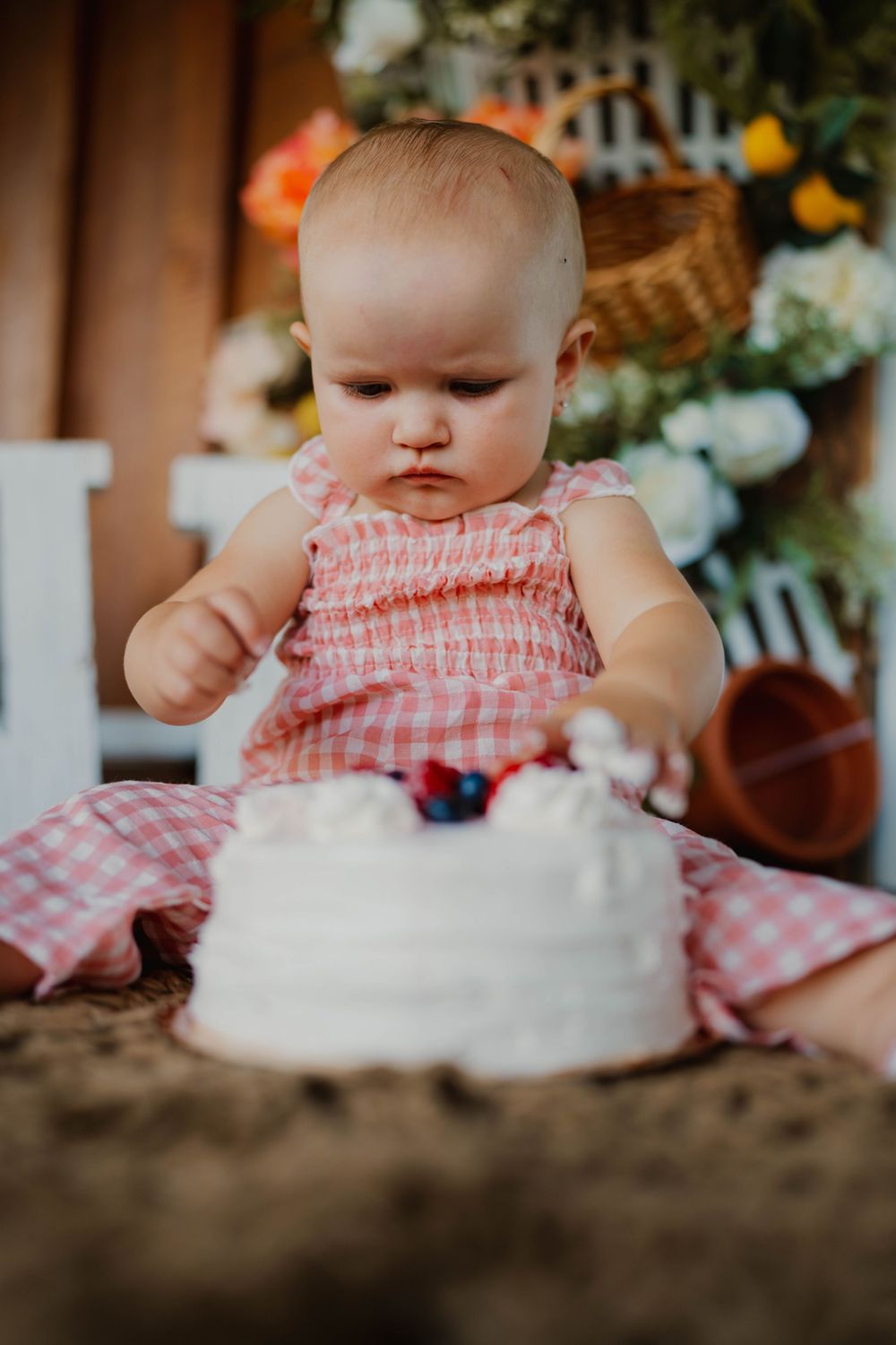 Baby in pink gingham dress reaching for white birthday cake surrounded by flowers.