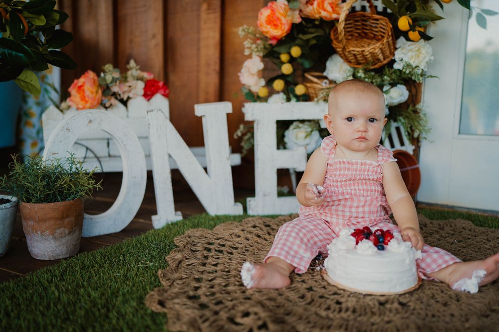 Baby in pink dress sitting by ONE sign and white cake for first birthday celebration.