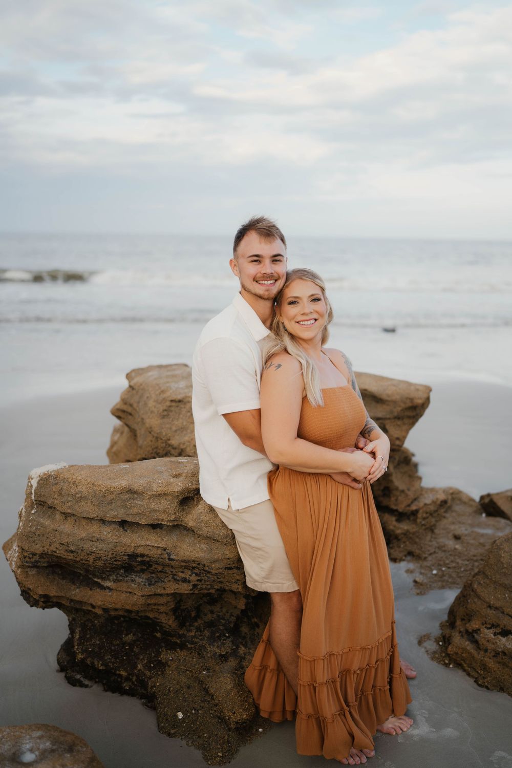 A couple embraces while posing on coastal rocks at sunset, with her wearing a flowing rust-colored dress.