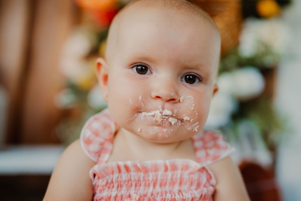 Close up of baby with cake frosting on face wearing pink gingham dress.
