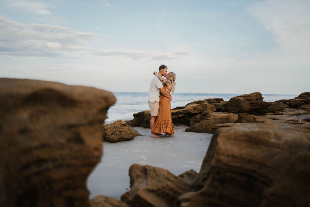 A romantic coastal photoshoot with two people standing among rocky tide pools at dusk.