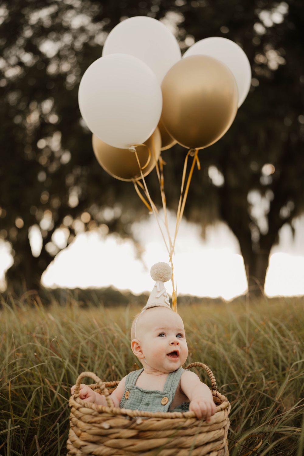 Baby sitting in woven basket with white balloons during golden hour outdoor photoshoot.