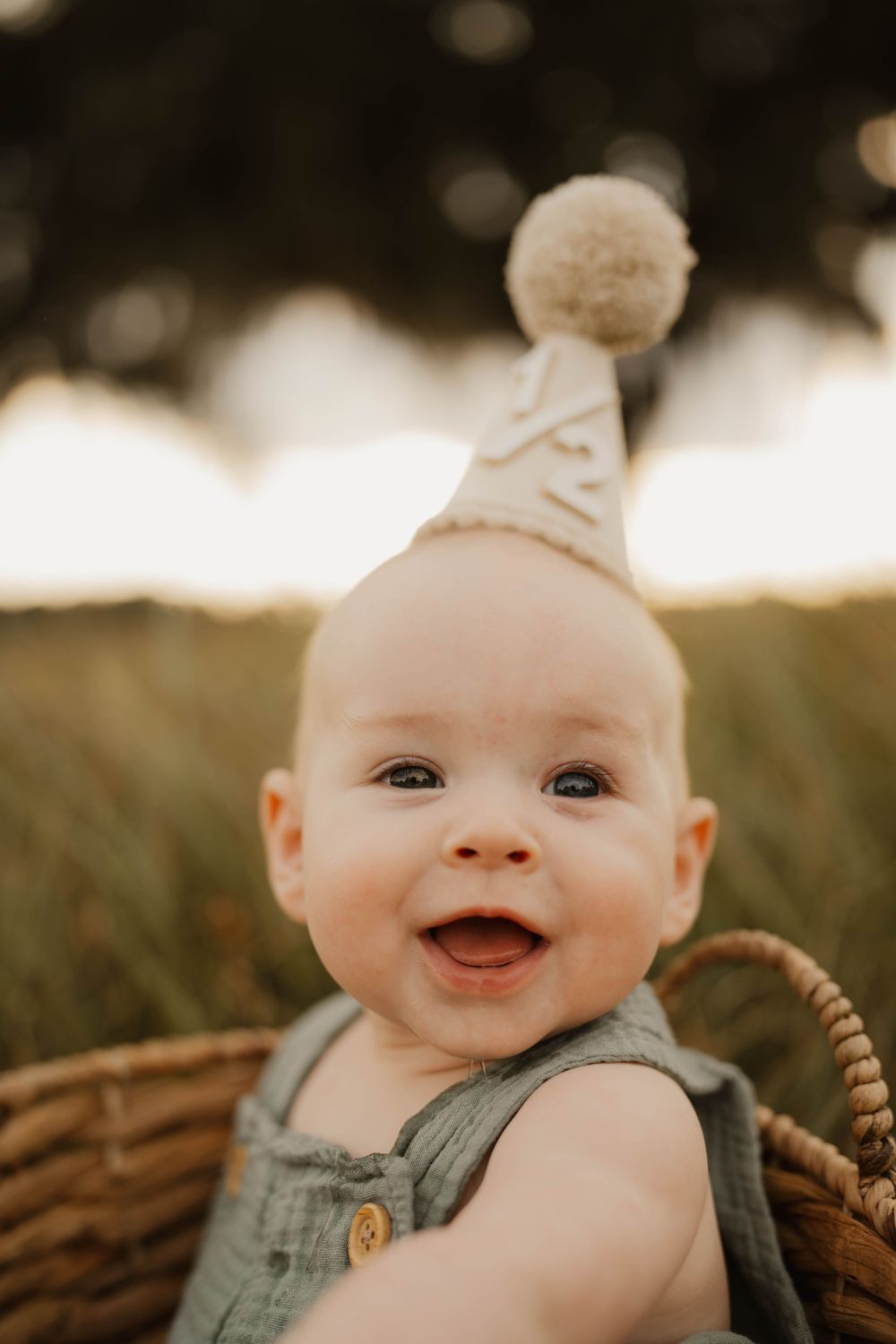 Happy baby wearing denim outfit with white party hat sitting in basket during sunset photoshoot.