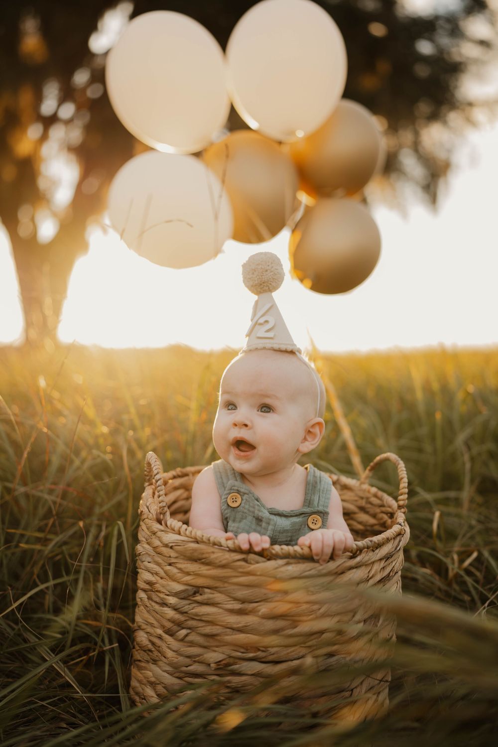 Baby wearing party hat sits in woven basket with white balloons during golden hour photoshoot.