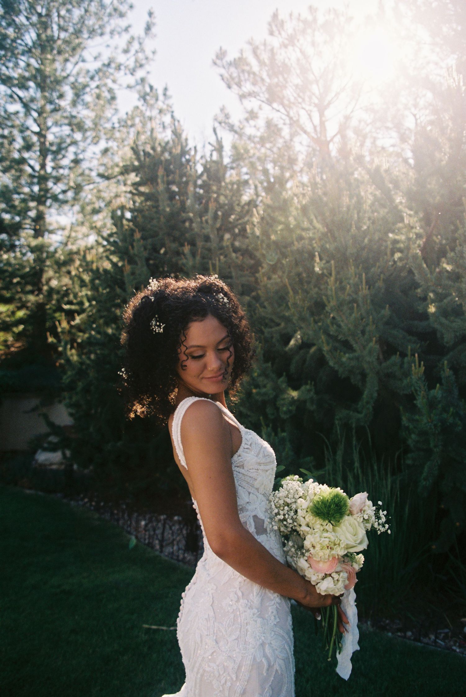 Beautiful bride in lace gown holding white floral bouquet poses in sunlit garden with evergreen trees in background.