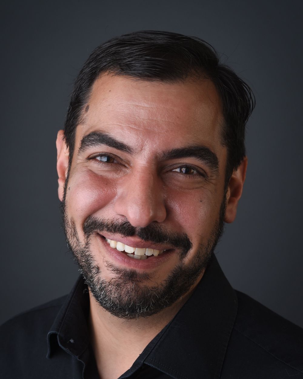 Headshot of a bearded man in a black shirt, smiling warmly, set against a dark grey backdrop at Abigail's Studio.