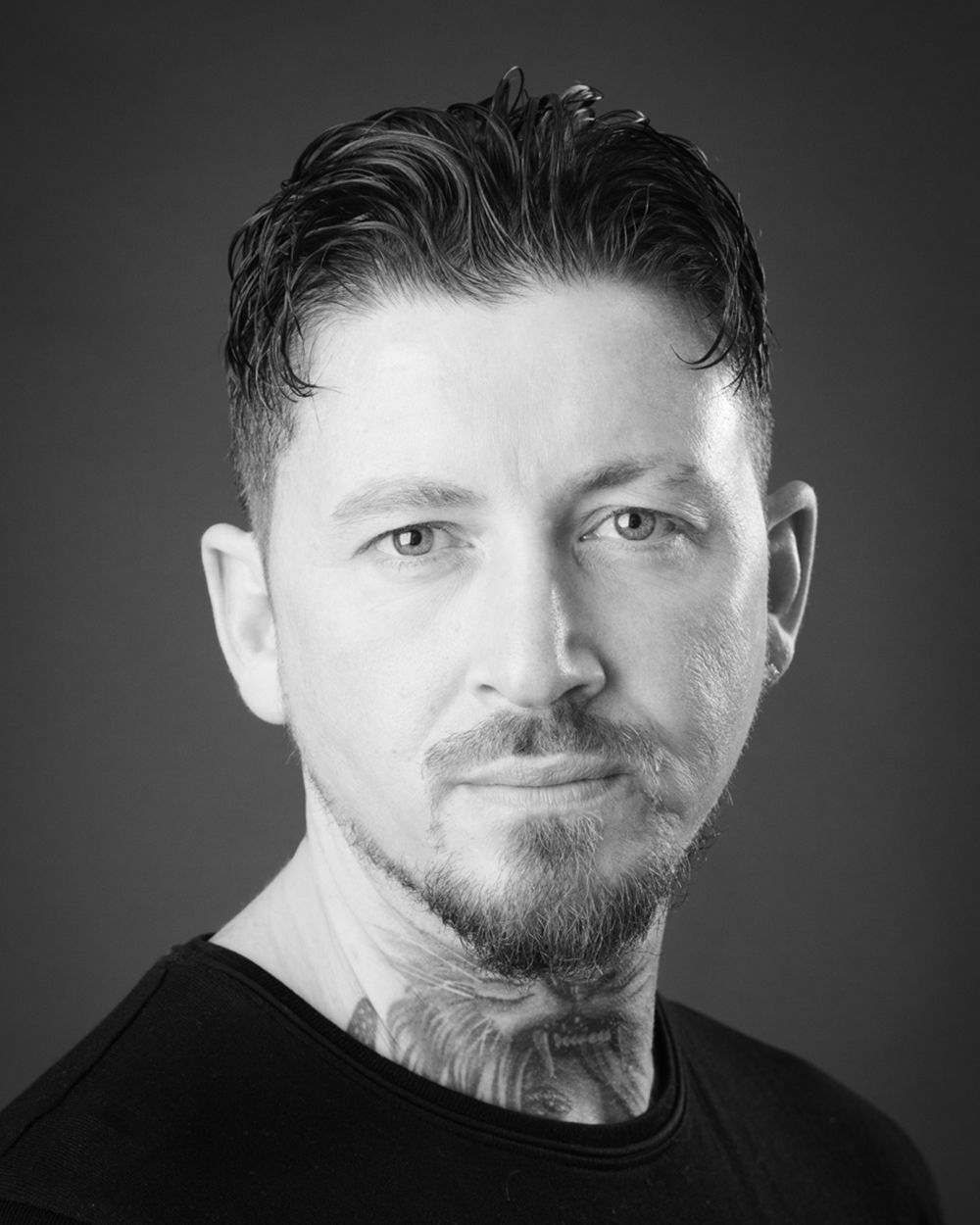 A man with tattoos wears a black shirt, captured in a dramatic pose against a dark grey backdrop at Abigail's Studio.