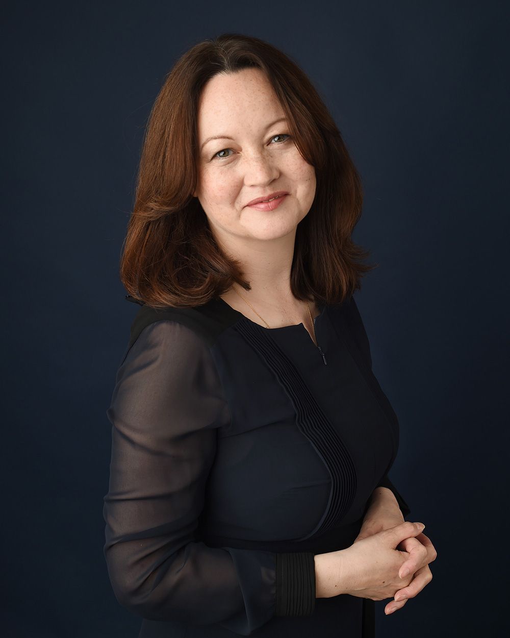 A woman with brown hair in a black dress poses for a LinkedIn headshot, featuring a dark blue background from Abigail's Studio.