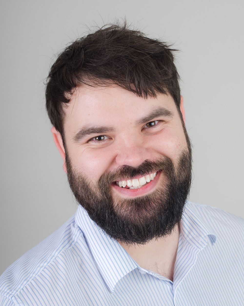A man with a beard and shirt smiles in a professional headshot, featuring a light grey background from Abigail's Studio.