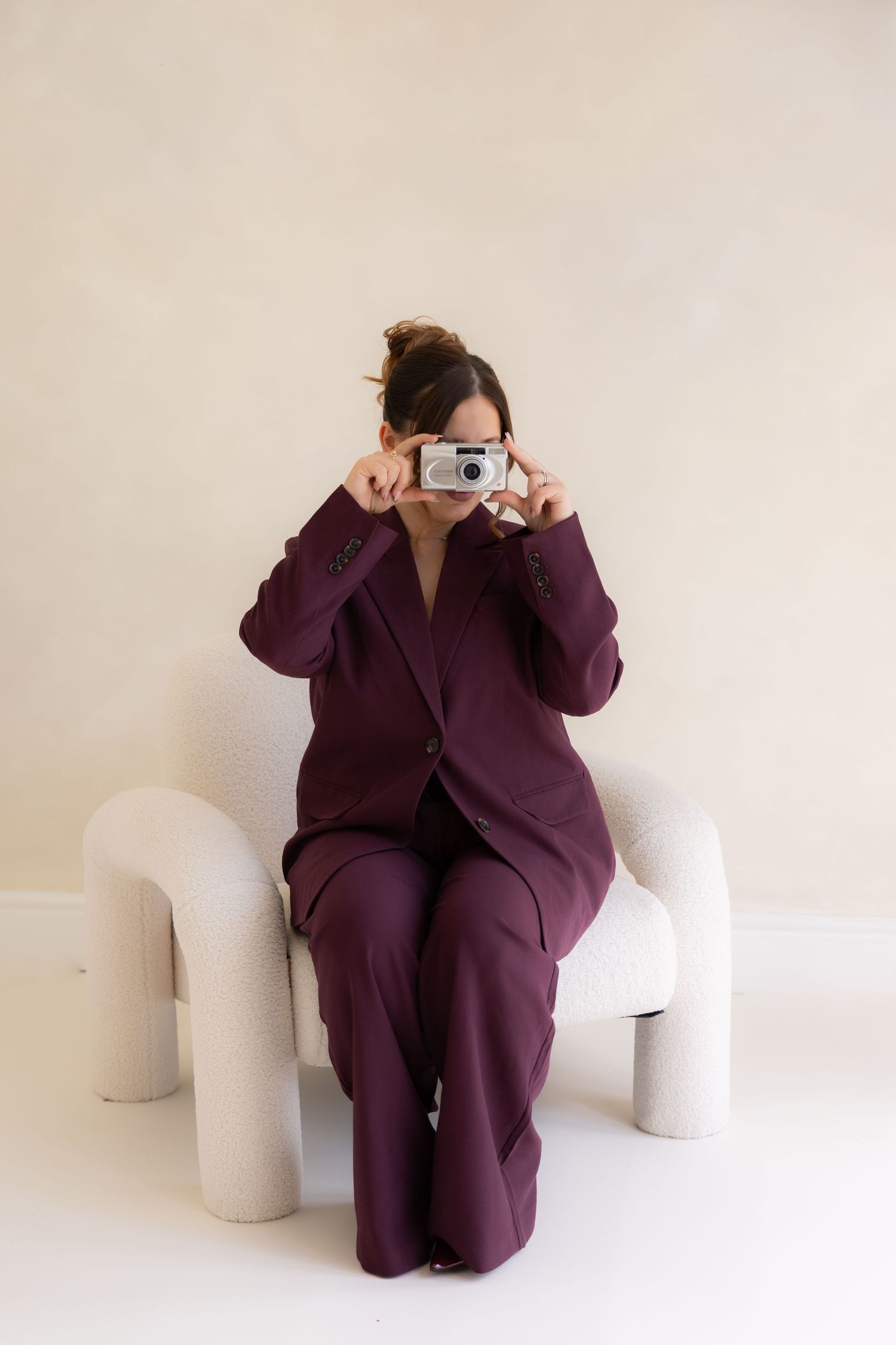 photographer holding 35mm film camera while sitting in boucle chair in a studio setting while wearing a burgundy suit.
