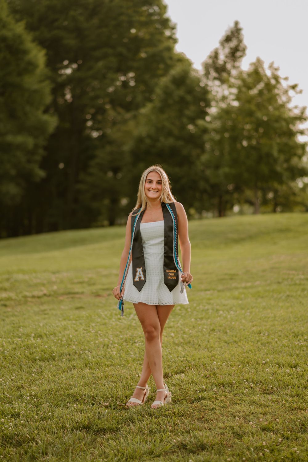 Maddy, a Blythe Photo senior client, wearing a white dress and graduation stole stands on a grassy field.