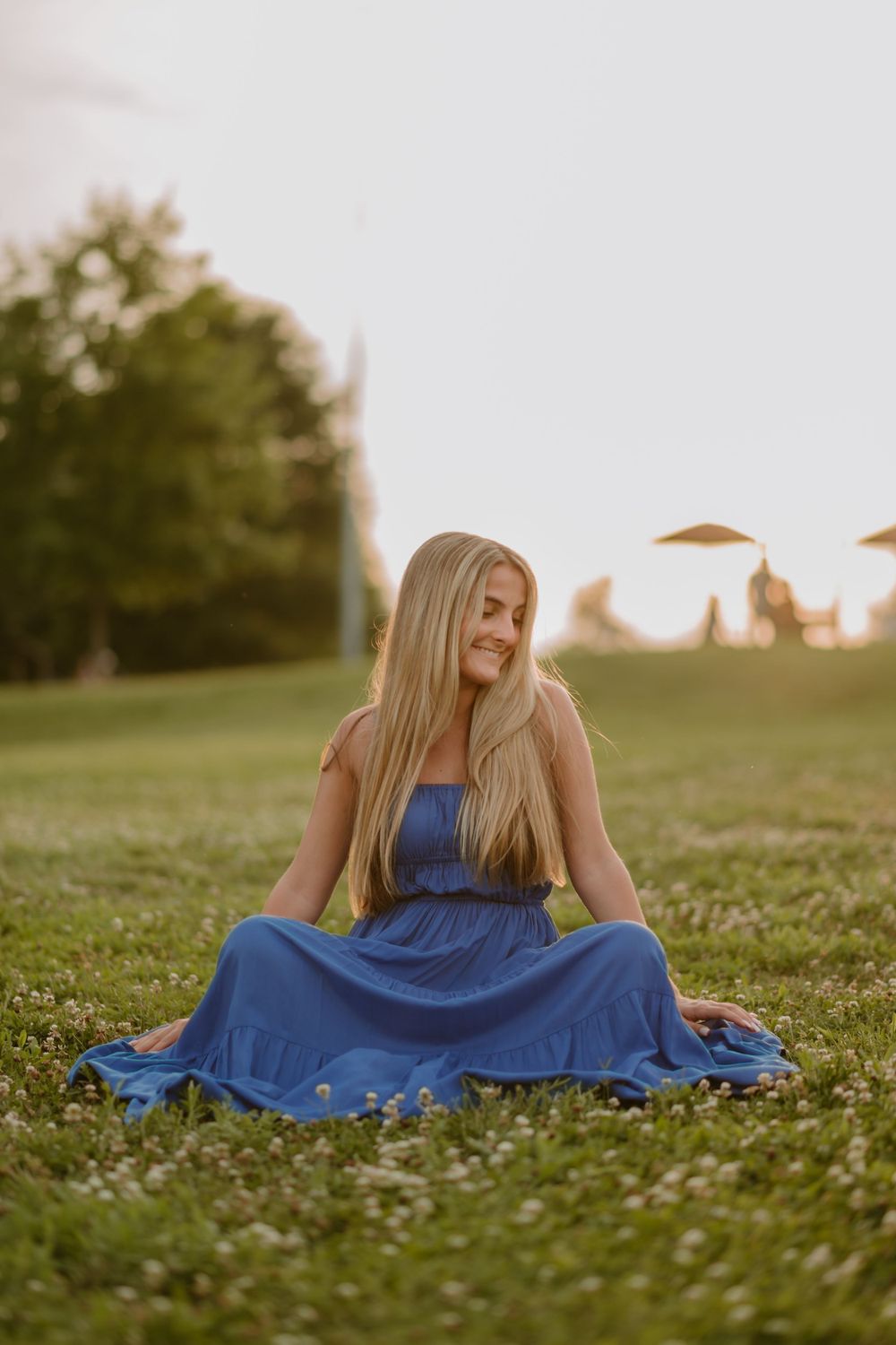 Woman in a blue dress sits on a grassy field during sunset, looking off to the side with a smile.