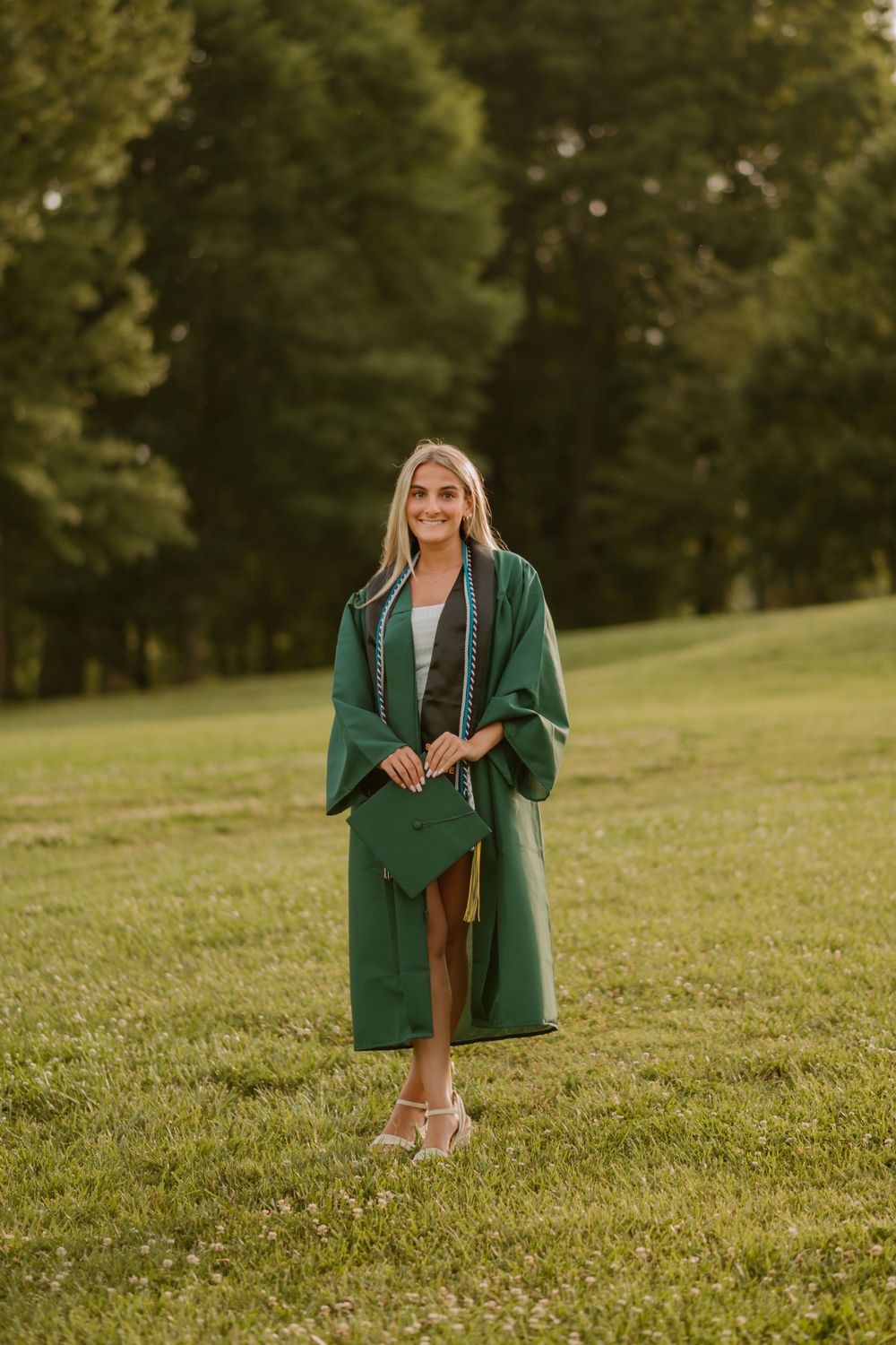A young woman in a white dress and graduation stole stands on a grassy field with trees in the background.