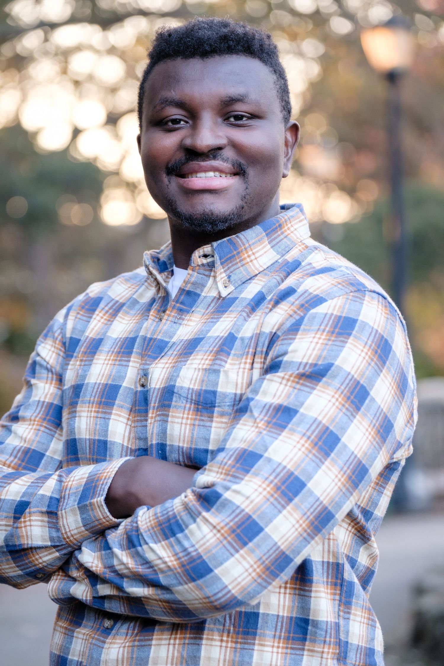 A person wearing a blue and white plaid shirt poses with crossed arms and a warm smile outdoors during golden hour.