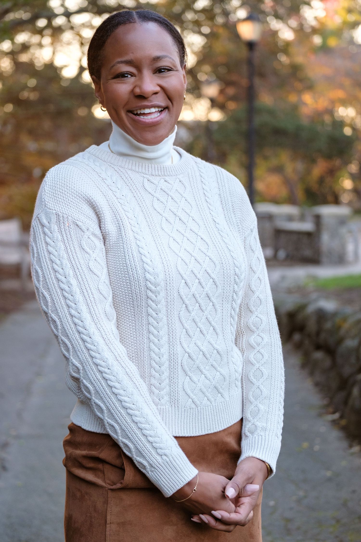 A person wearing a white cable knit turtleneck sweater and brown skirt stands outdoors in an autumn setting.