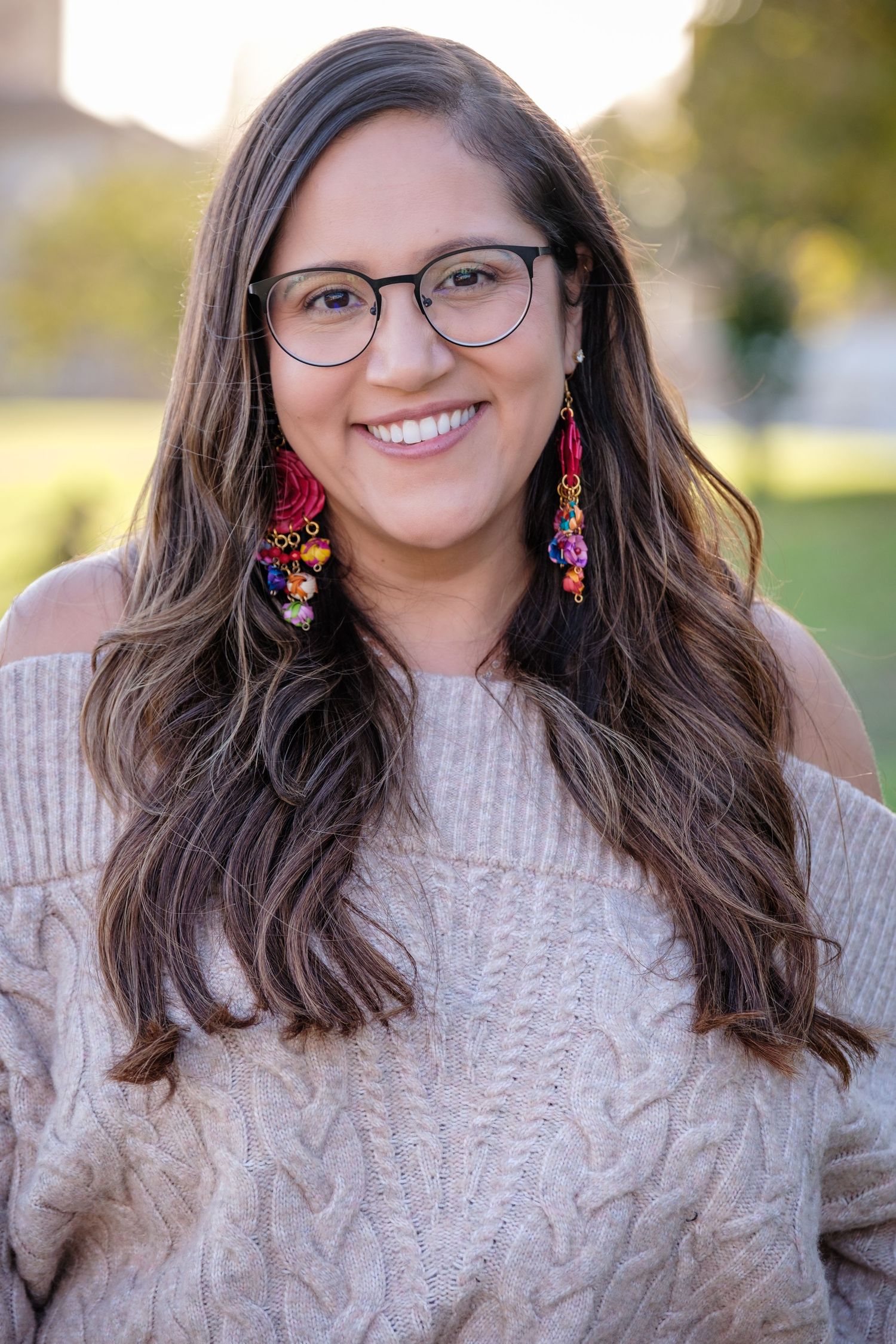 Smiling young woman with long brown hair, glasses, and colorful tassel earrings wearing a cozy beige cable-knit sweater outdoors.