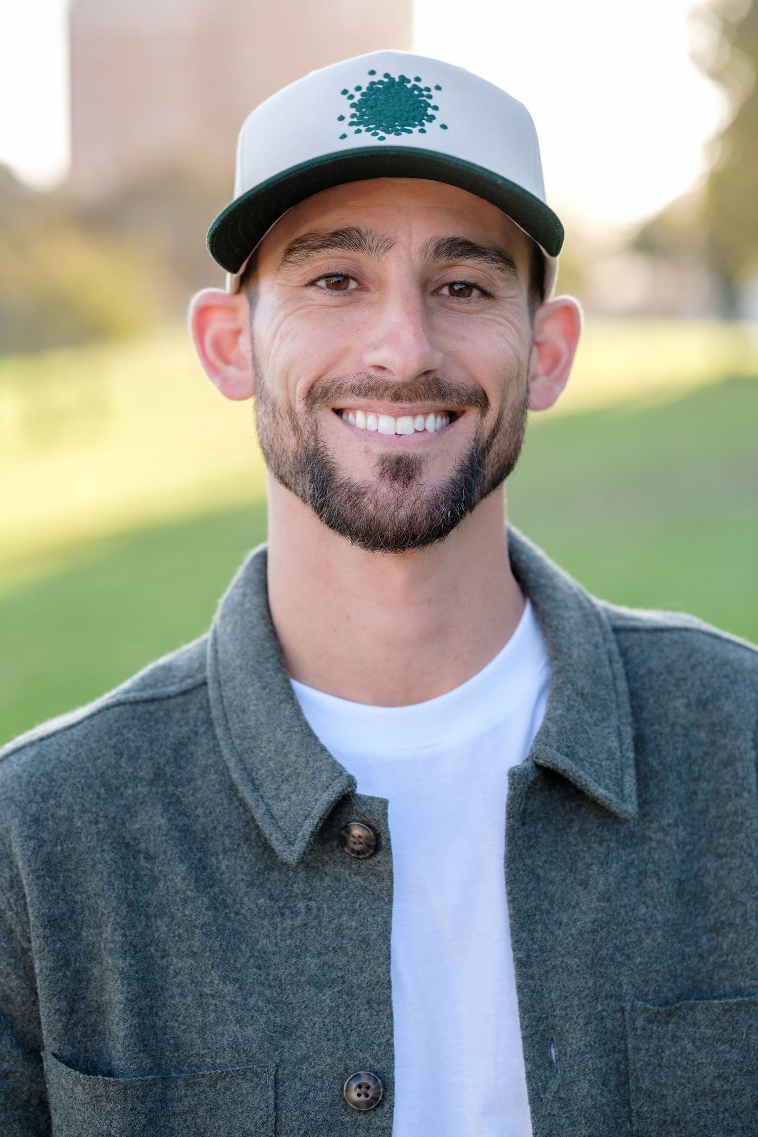 Smiling young man with beard wearing a green cap and gray jacket outdoors in a park setting.