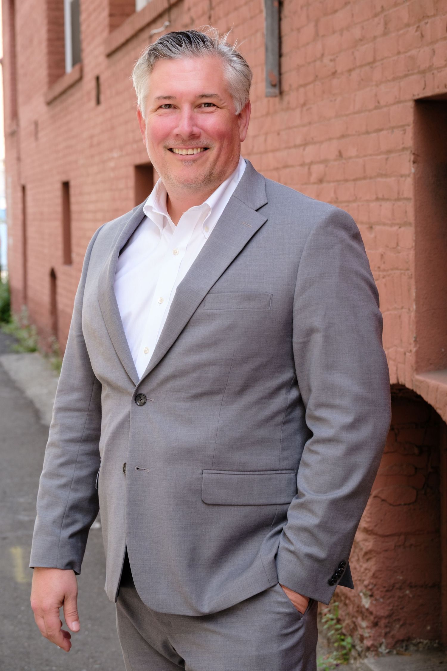 Smiling middle-aged man in gray suit and white shirt posing outdoors near a red brick building.