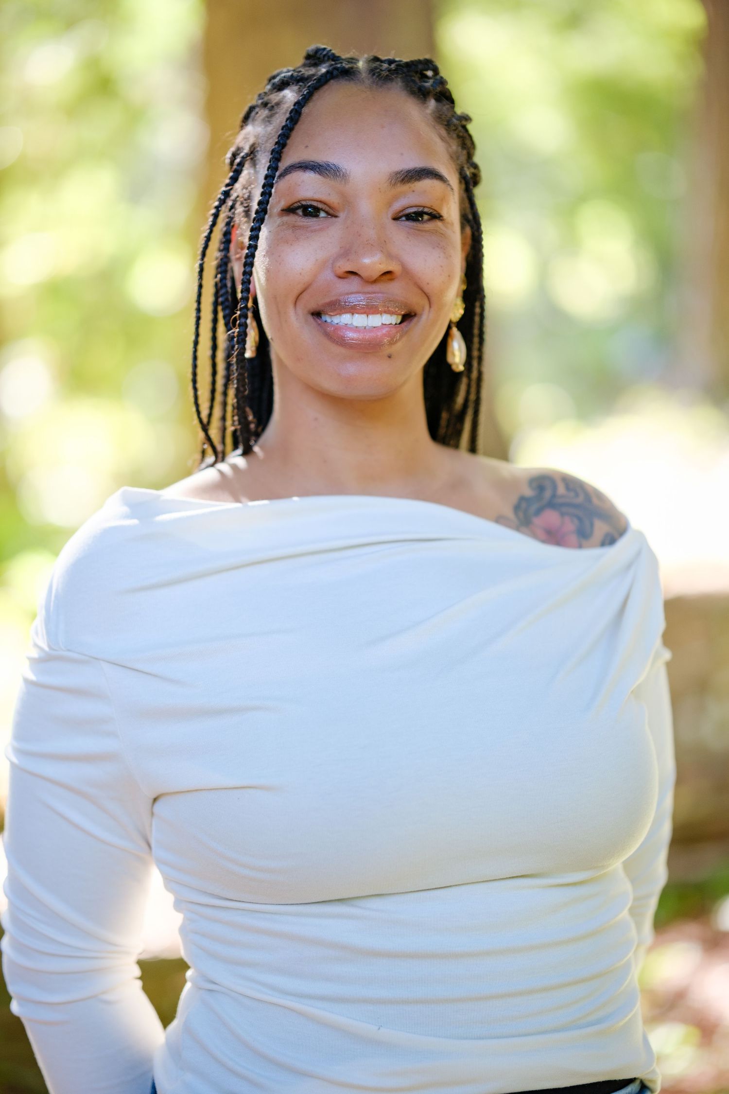 Smiling woman with braids wearing white off-shoulder top, tattoo visible, standing outdoors in natural light.