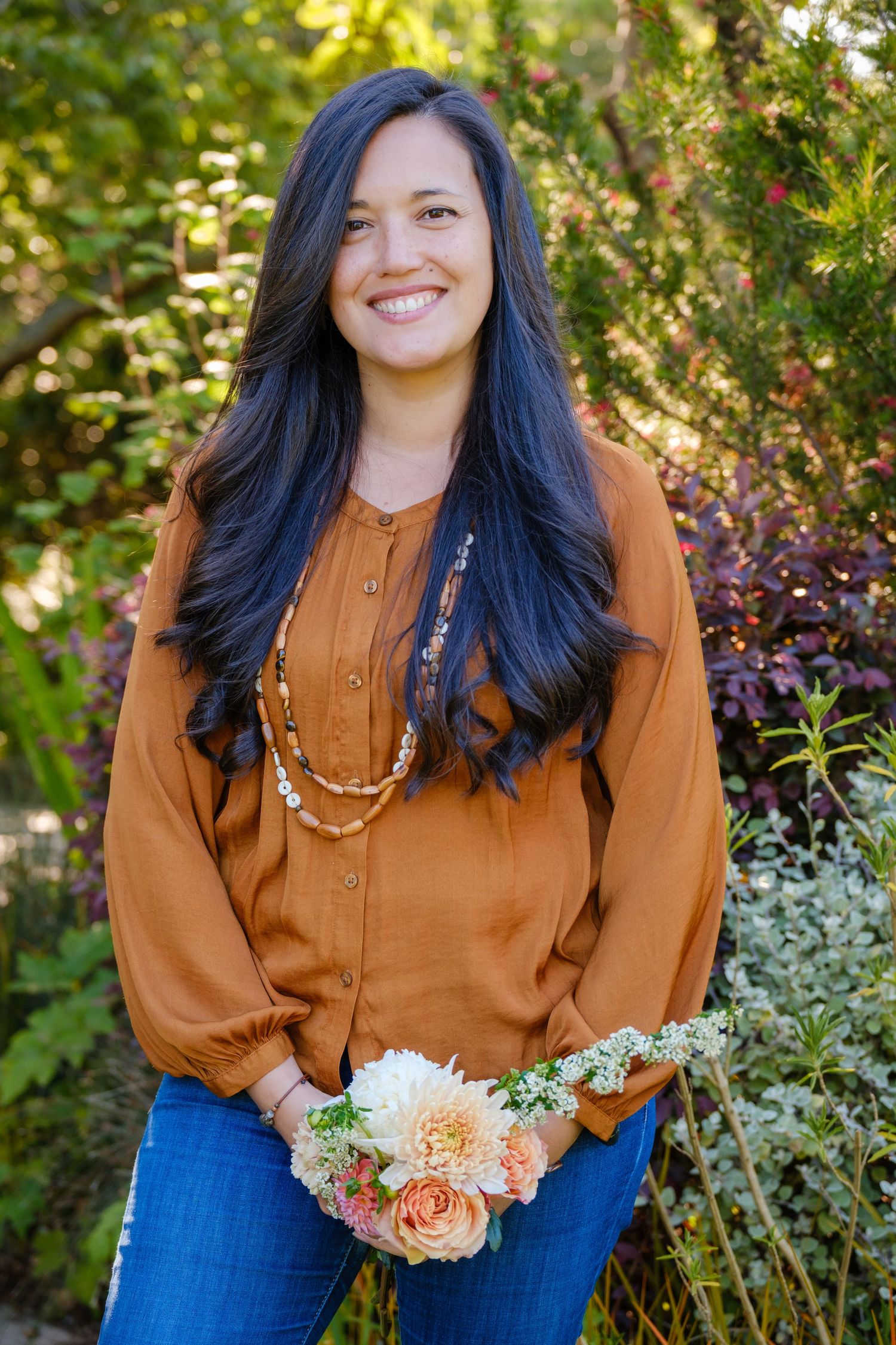 Smiling woman with long dark hair wearing a rust orange blouse and jeans, holding peach flowers in a lush garden.