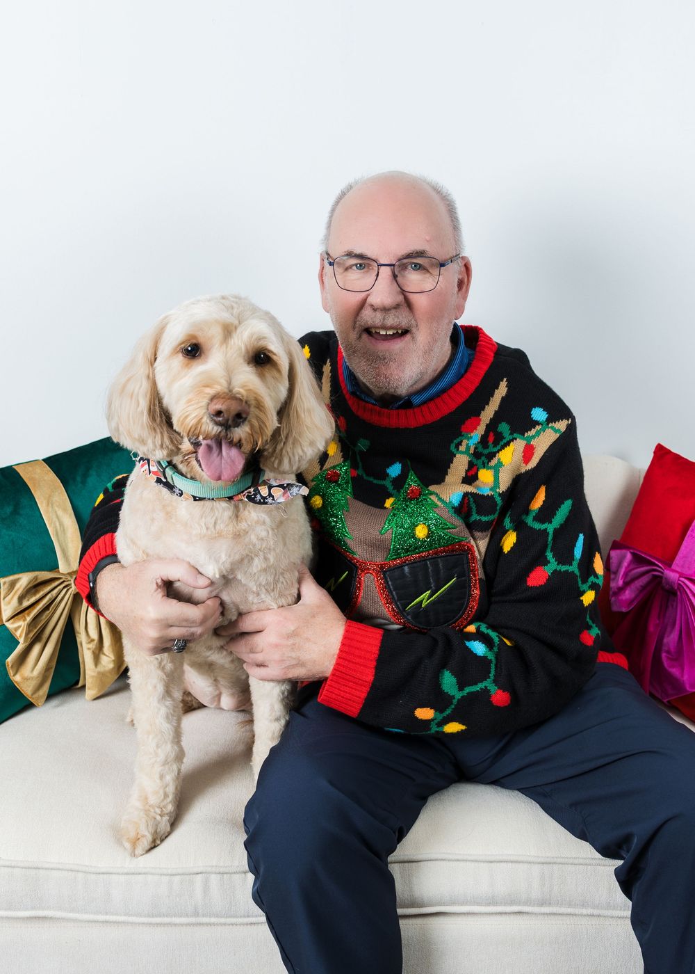 A smiling bald man in a Christmas sweater poses with his fluffy golden doodle dog on a white couch.
