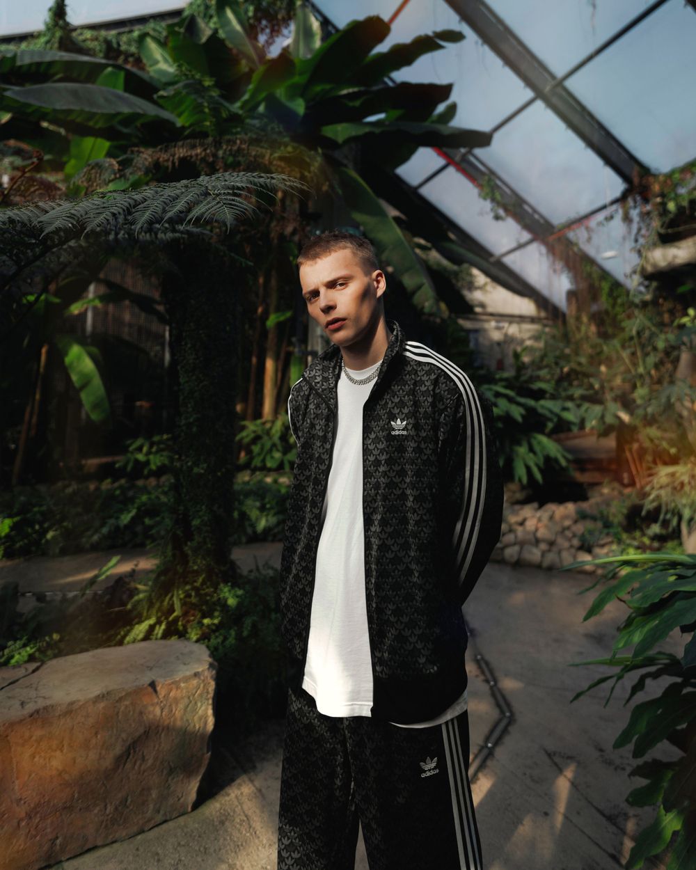 Lifestyle portrait of a man in a dark Adidas jacket standing among tropical plants in a glass greenhouse