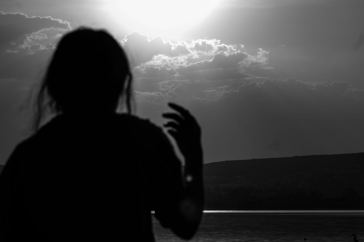 Silhouette of a person raising their hand against a dramatic sunset sky over water in black and white.