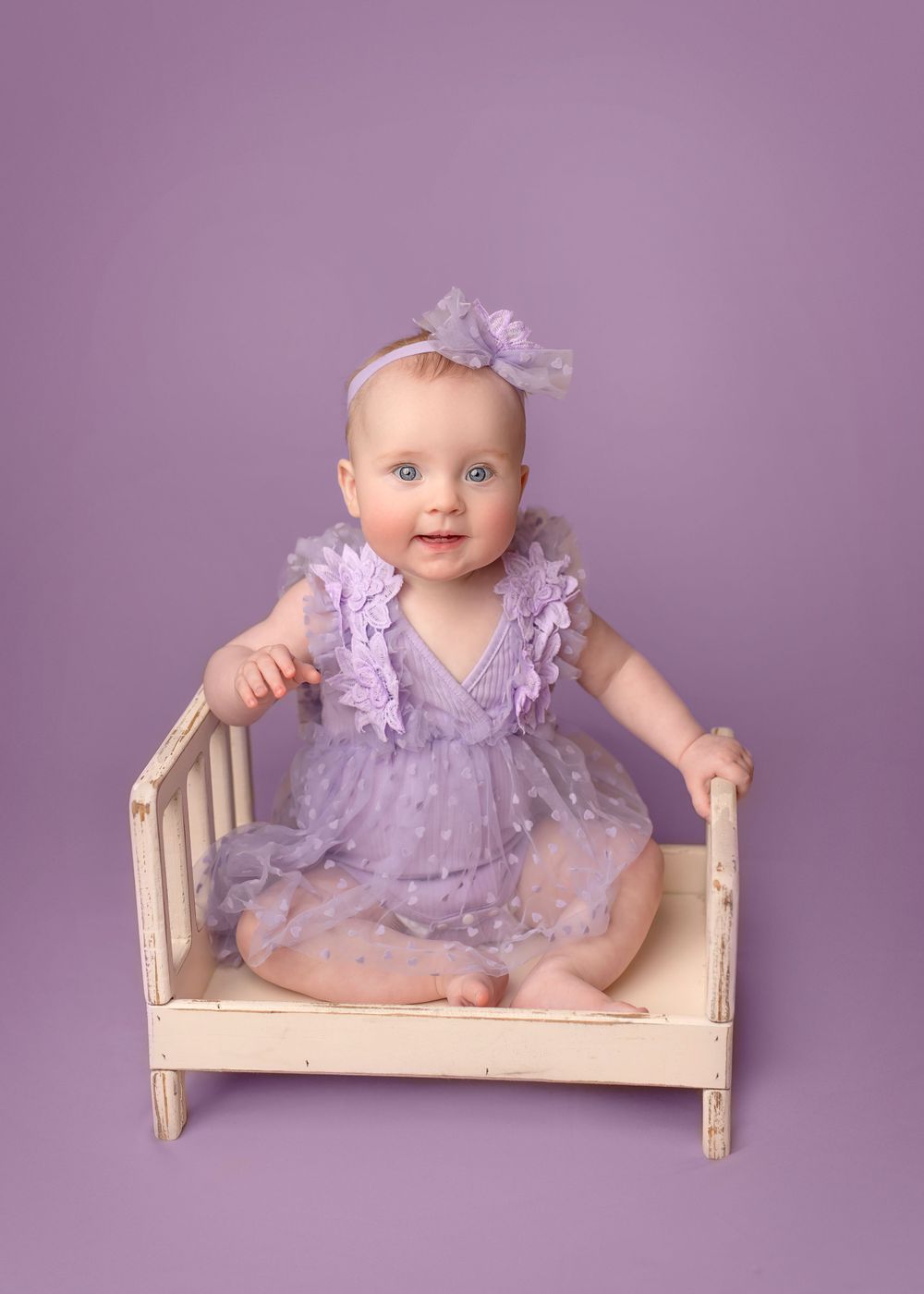 A baby in a pale purple dress with ruffles and matching headband sits in a small whitewashed wooden bench against purple backdrop. Sitter.