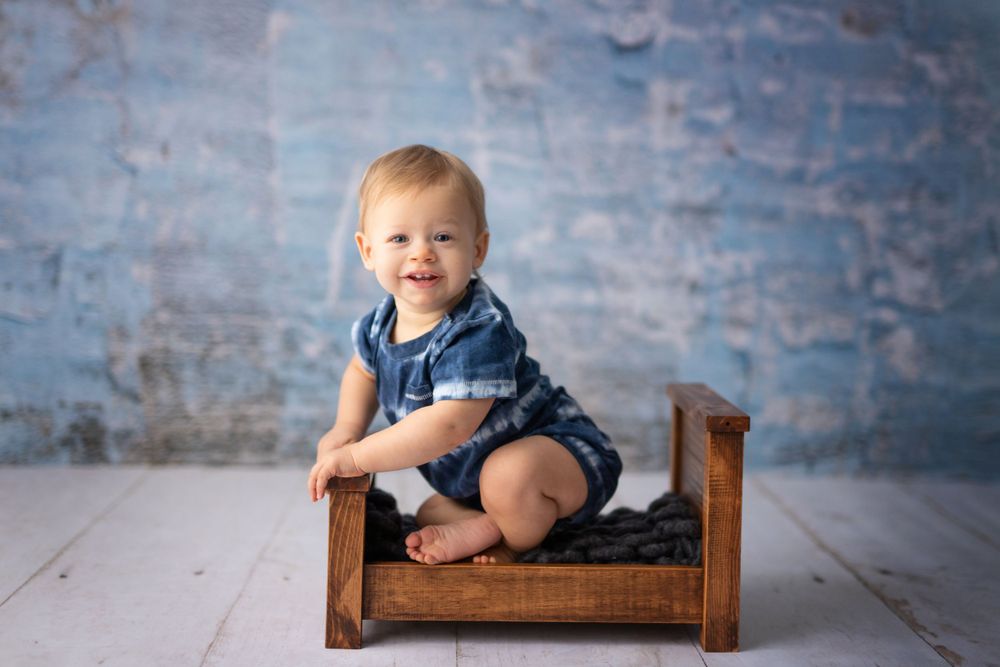 A toddler in navy blue overalls sits and smiles while posing on a small wooden bed against a blue rustic backdrop. Sitter Session.