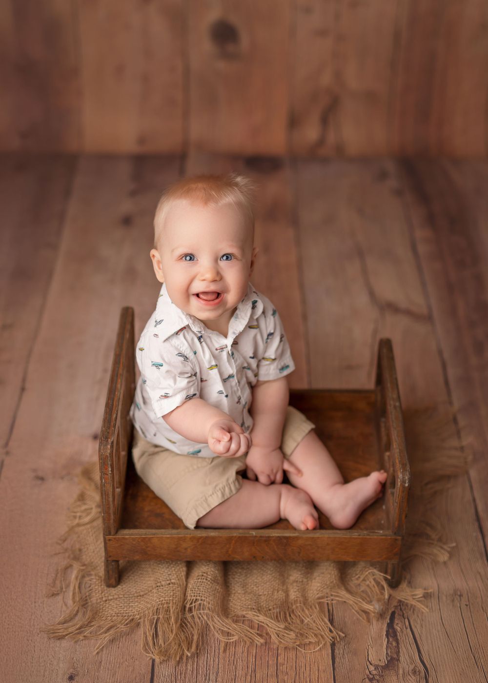A smiling baby wearing a light colored shirt and khaki shorts sits in a vintage wooden crate on a wooden floor. Milestone session.