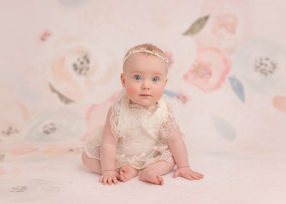 Small baby in white lace outfit sits on floral backdrop in soft pastel photography studio setting.