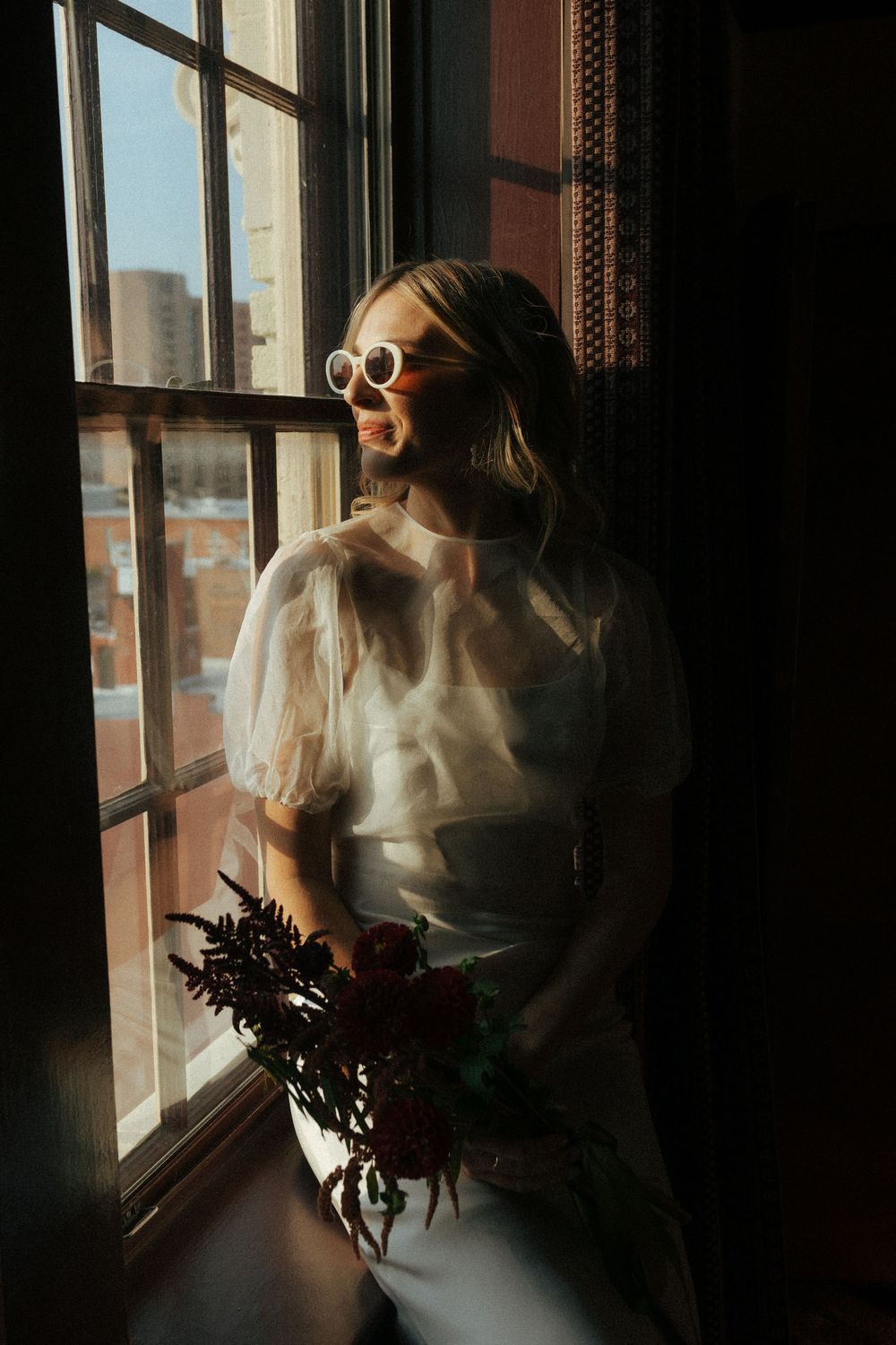 A dreamy bridal portrait by a window with sunlight streaming in, featuring a white dress and sunglasses.
