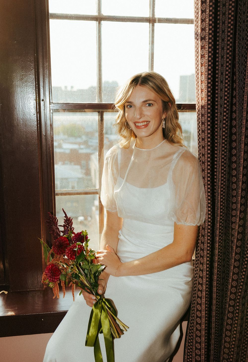 Bride in elegant white dress with sheer sleeves poses by window holding burgundy bouquet.