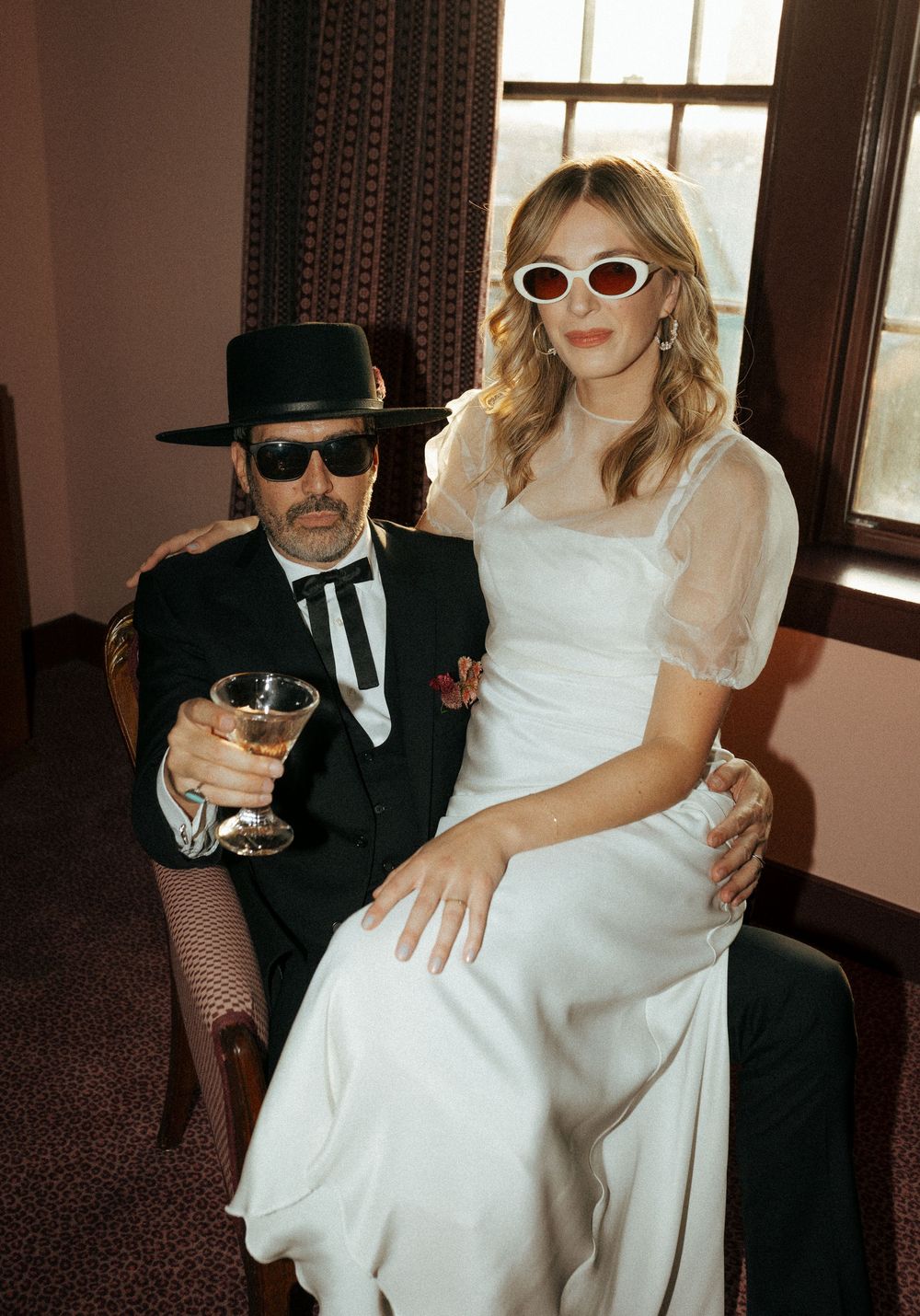 Wedding couple in vintage-inspired attire with sunglasses and hat pose for stylish portrait.