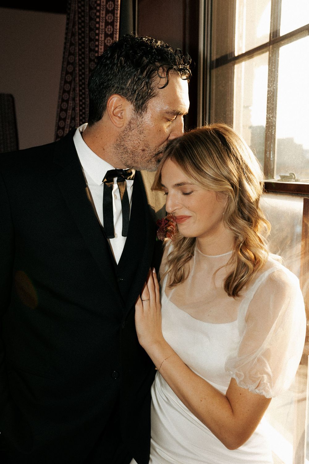 Romantic couple in formal attire sharing intimate moments by sunlit window.
