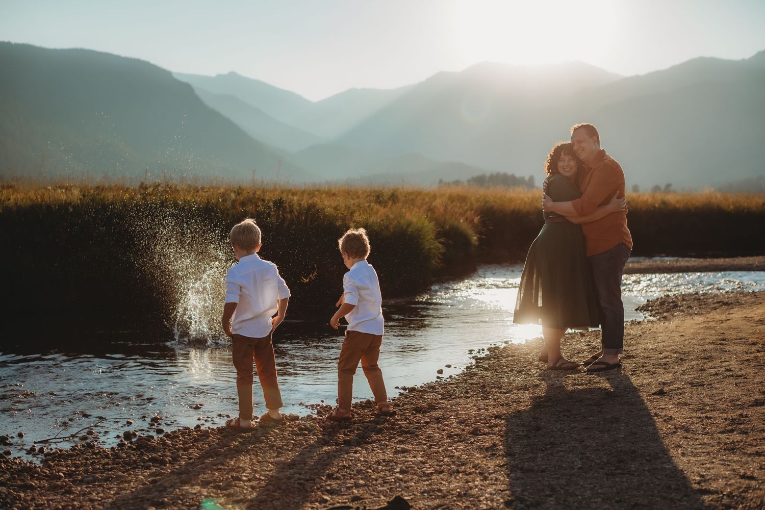 Family silhouetted against sunset by mountain lake with children playing at water's edge.